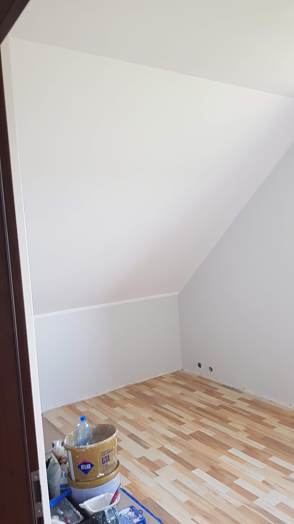 Poddasze po malowaniu na biało, widoczne narzędzia malarskie i wiadro z farbą Atlas GTA. Podłoga wyłożona panelami imitującymi drewno. Świeżo pomalowane ściany.
