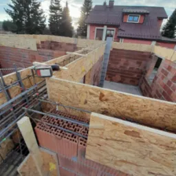 Budowa domu w trakcie: mury z cegły, szalunki z płyty OSB, zbrojenie. W tle dom z czerwonym dachem i zielone drzewa pod błękitnym niebem. Widok z góry.