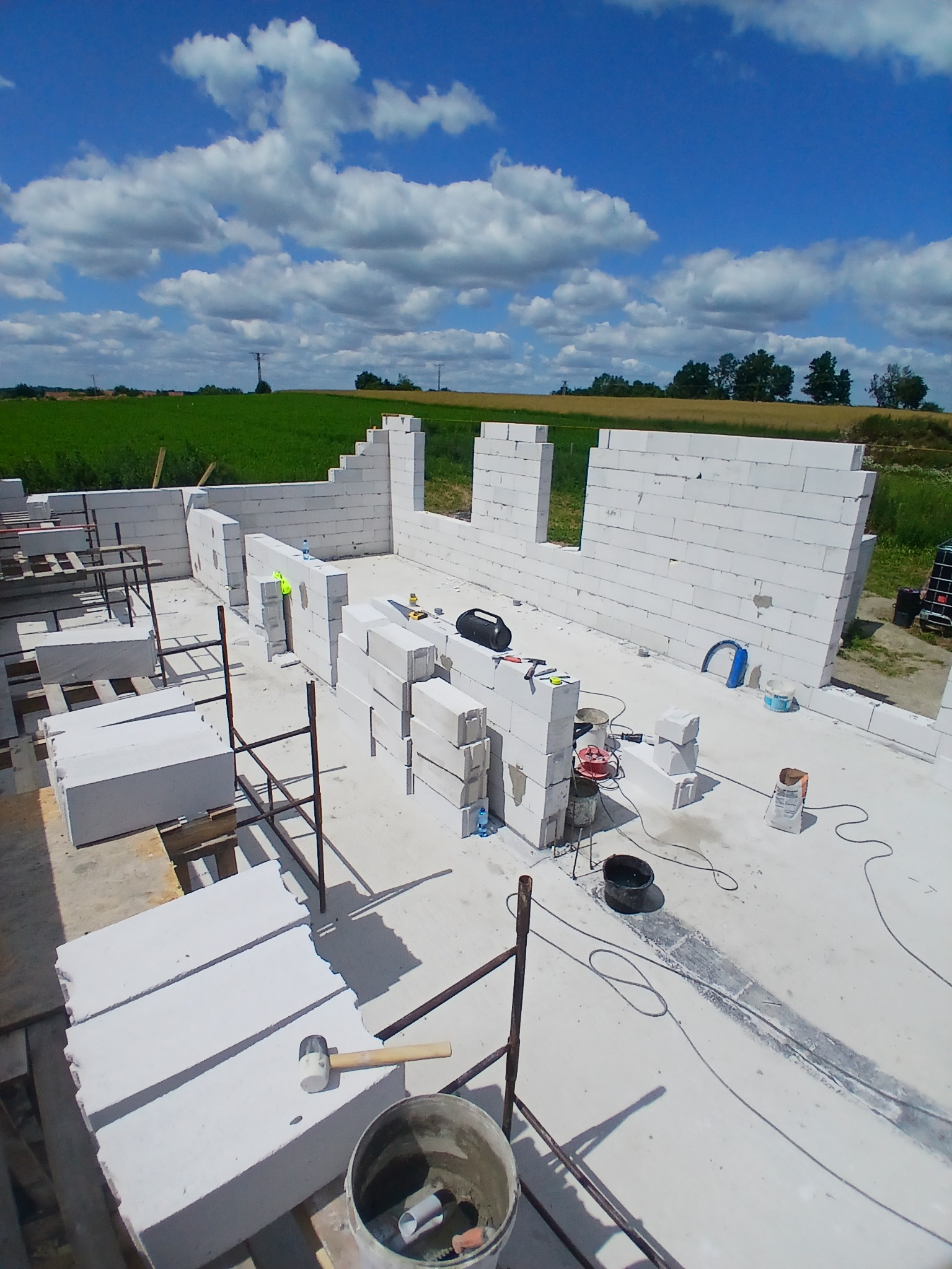 Wznoszenie ścian domu z białych bloczków, widok z góry na plac budowy w słoneczny dzień. Częściowo postawione ściany, narzędzia i materiały budowlane.