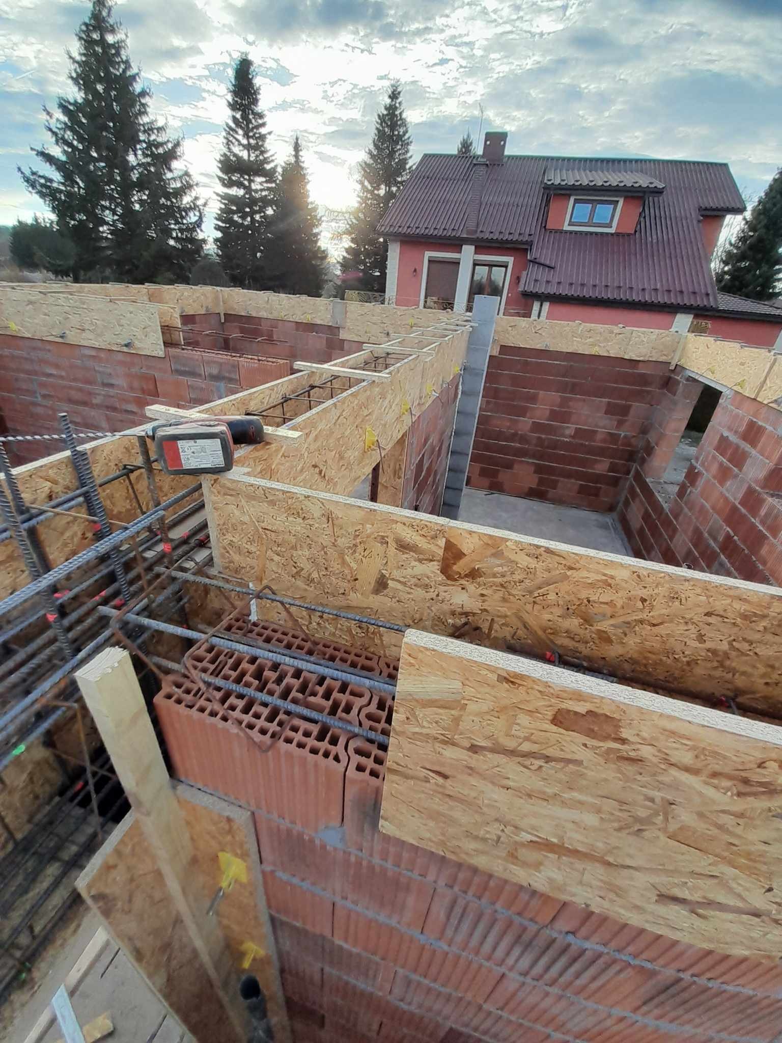 Budowa domu w trakcie: mury z cegły, szalunki z płyty OSB, zbrojenie. W tle dom z czerwonym dachem i zielone drzewa pod błękitnym niebem. Widok z góry.