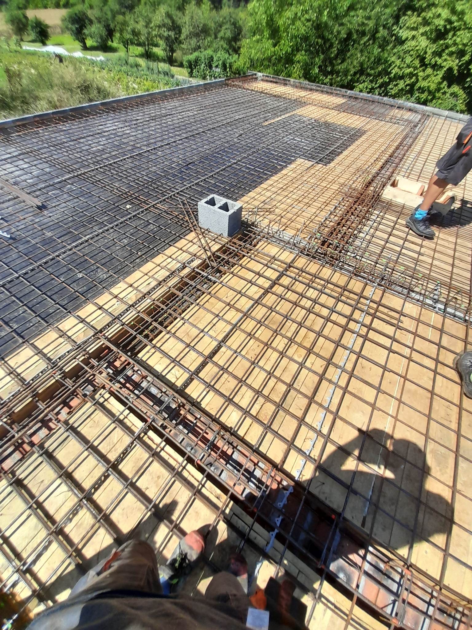 Zbrojenie stropu na deskowaniu z widocznymi prętami, siatką zbrojeniową i częściowo ułożoną folią. W tle zieleń drzew. Kąt ujęcia z góry.