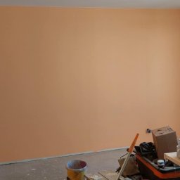 Sebastian Matwiejuk - Pomarańczowa ściana po malowaniu w pokoju z widocznymi narzędziami malarskimi, otwartym balkonem i jasnym oświetleniem.