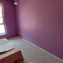 Sebastian Matwiejuk - Pomieszczenie po malowaniu ścian na fioletowo. Widoczne zabezpieczone okno, podłoga z plamami farby i drewniany stołek malarski.