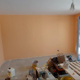 Sebastian Matwiejuk - Ściana pomalowana na brzoskwiniowy kolor w pokoju z otwartymi drzwiami balkonowymi. Widoczne narzędzia malarskie i niezabezpieczona podłoga.