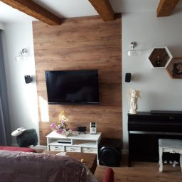 Sebastian Matwiejuk - Salon z panelami winylowymi na ścianie, świeżo szpachlowanymi i pomalowanymi ścianami oraz sufitem. Widoczne drewniane belki stropowe i dekoracje ścienne.