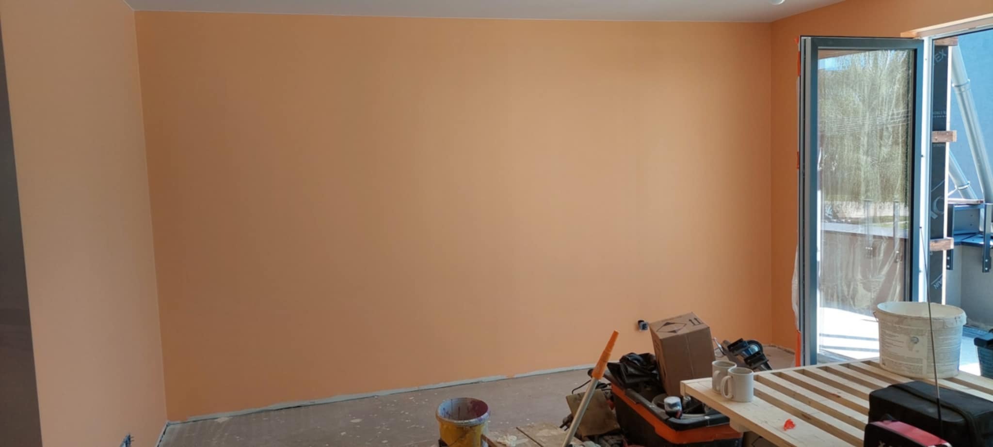 Pomarańczowa ściana po malowaniu w pokoju z widocznymi narzędziami malarskimi, otwartym balkonem i jasnym oświetleniem.