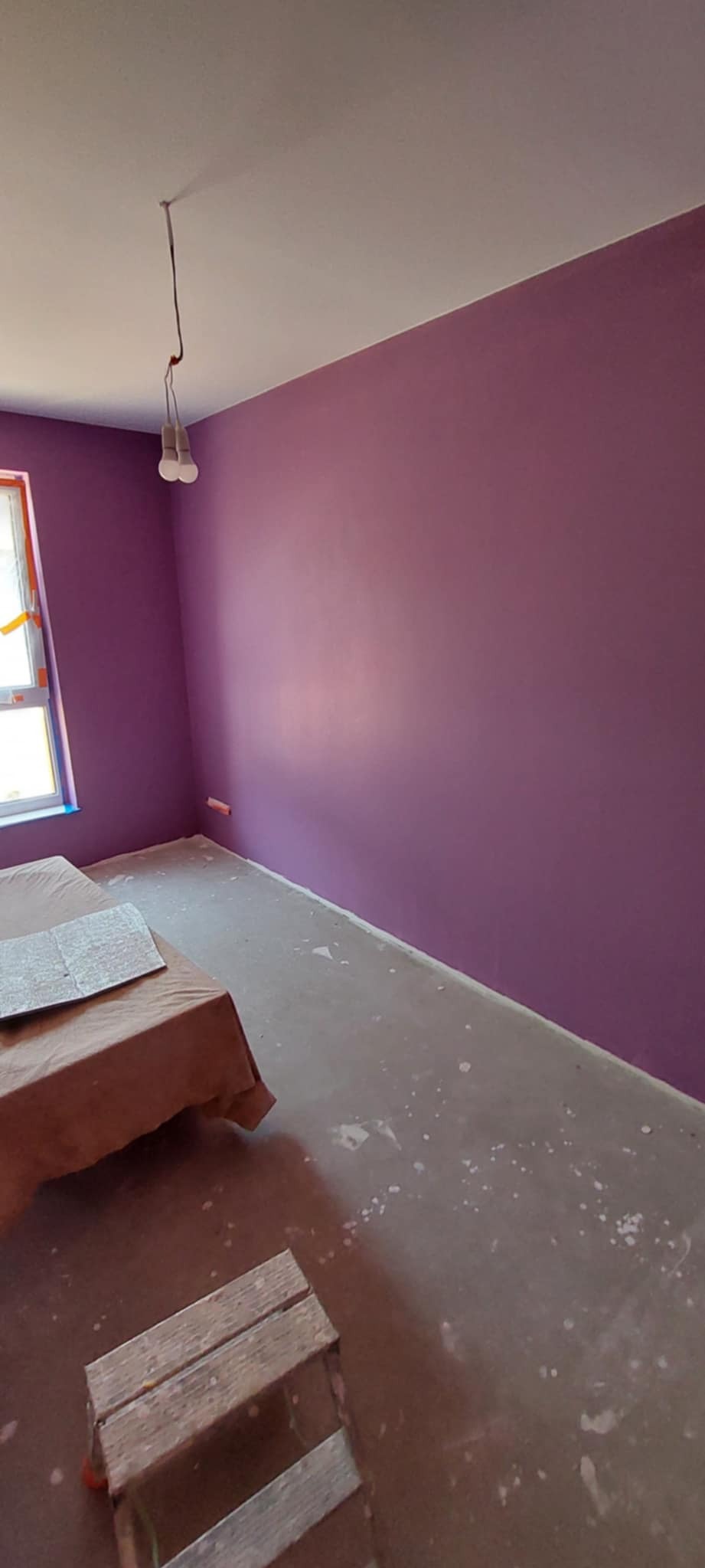 Pomieszczenie po malowaniu ścian na fioletowo. Widoczne zabezpieczone okno, podłoga z plamami farby i drewniany stołek malarski.