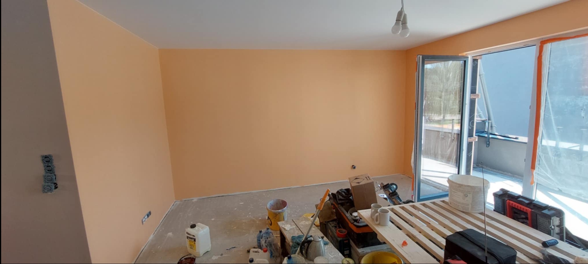 Ściana pomalowana na brzoskwiniowy kolor w pokoju z otwartymi drzwiami balkonowymi. Widoczne narzędzia malarskie i niezabezpieczona podłoga.