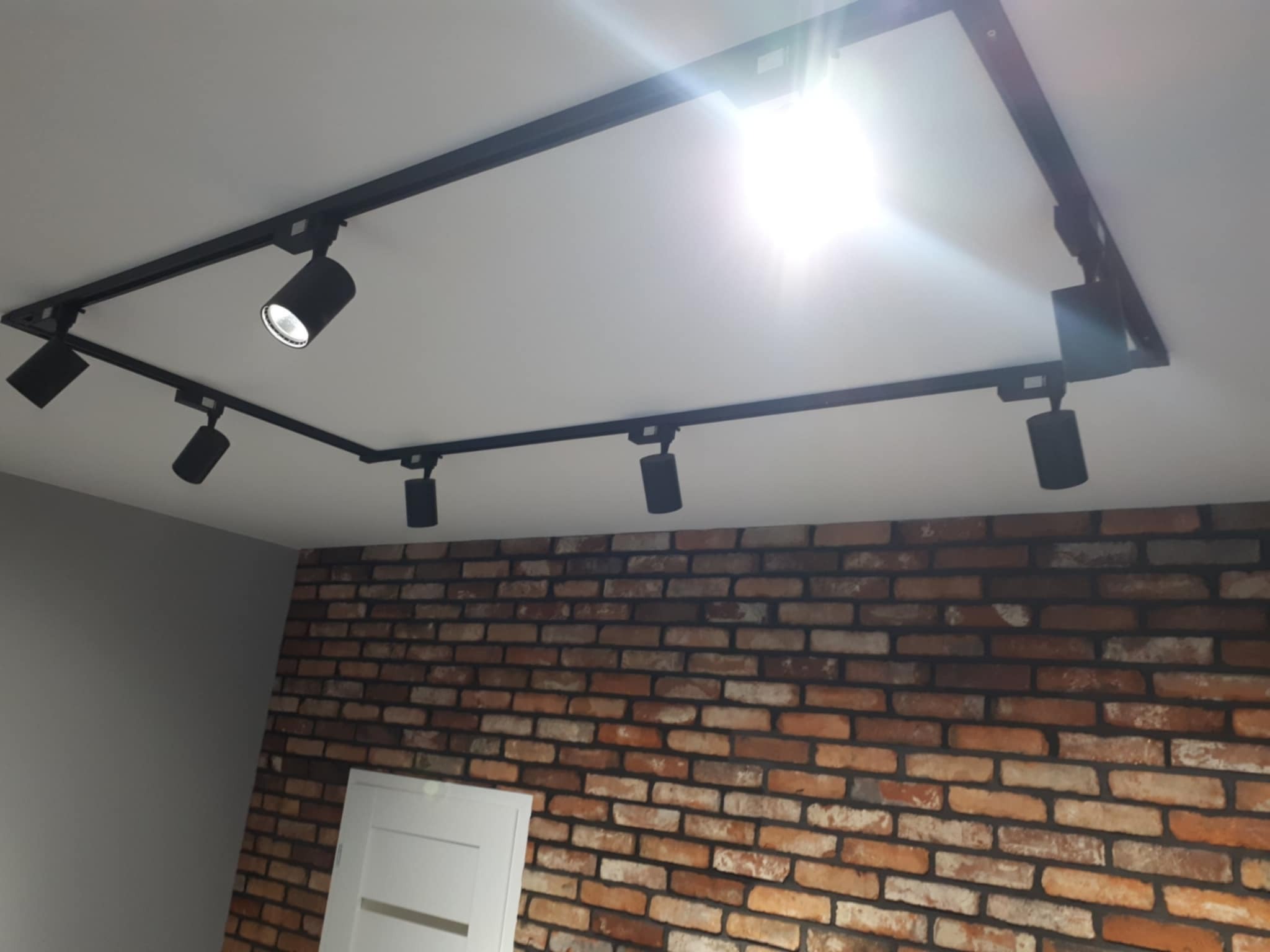 Ceglana ściana z oświetleniem szynowym w nowoczesnym wnętrzu. Część ściany gładka, szara. Widoczny fragment białych drzwi.