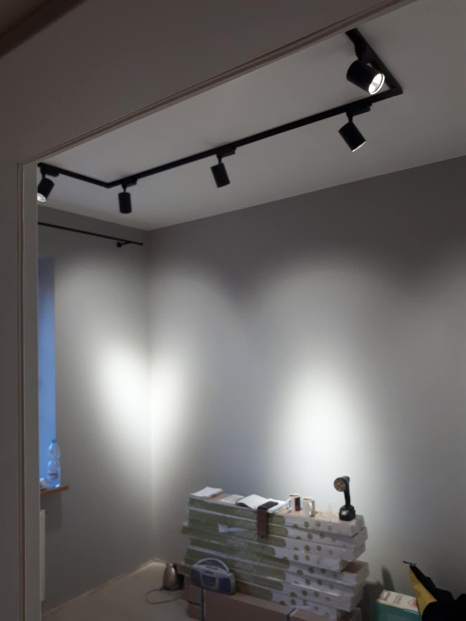 Szpachlowana ściana z oświetleniem szynowym. Na podłodze leżą materiały budowlane i narzędzia. Widok na świeżo wykończone wnętrze z nowoczesnym oświetleniem.