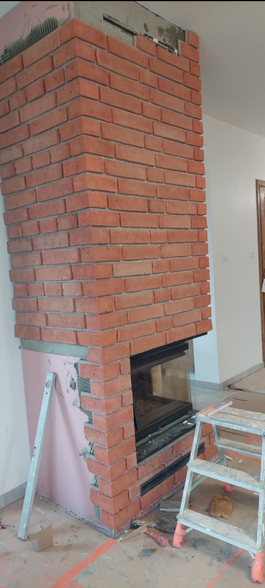 Wznoszenie kominka z cegły klinkierowej, widoczne narzędzia i drabina. Prace wykończeniowe w trakcie realizacji, surowy stan.