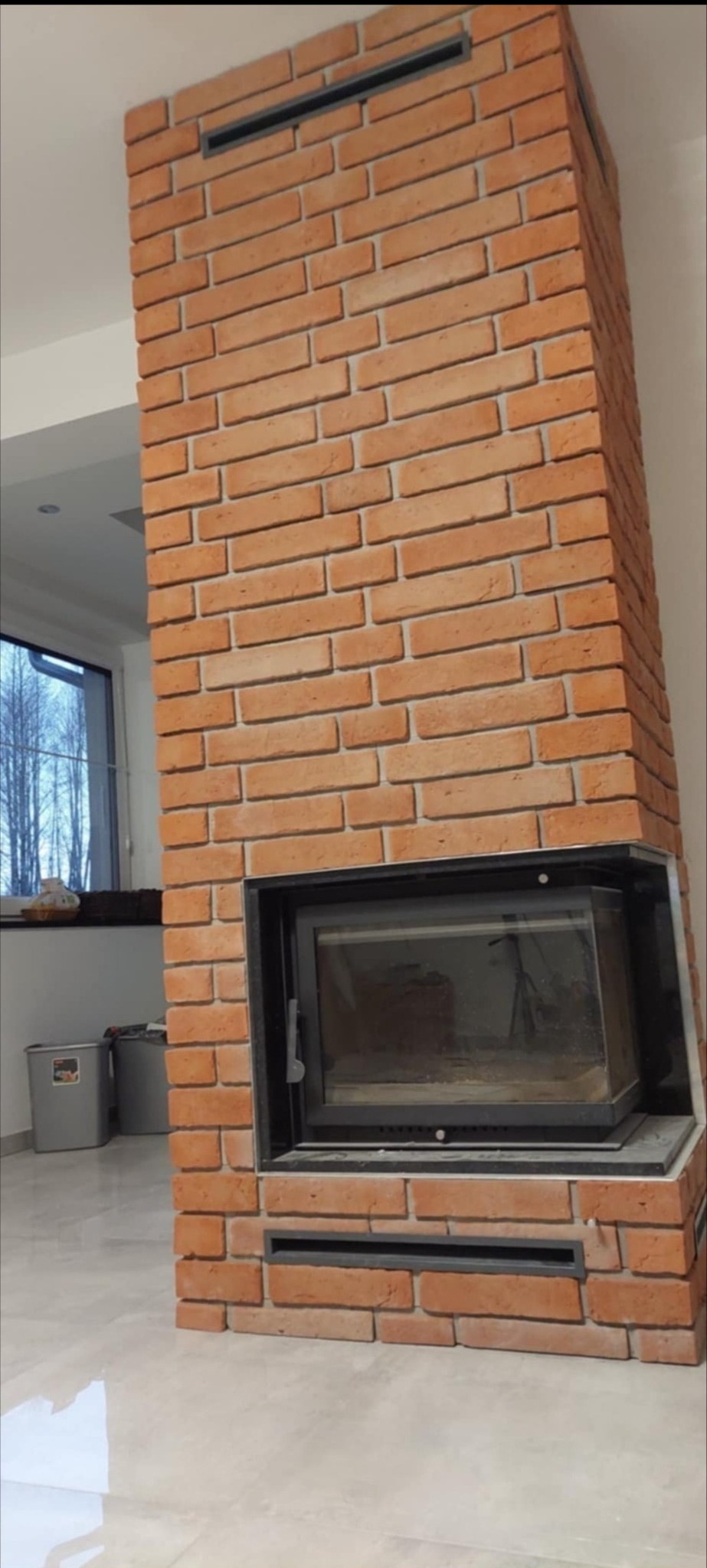 Murowany kominek z cegły w salonie, z czarną ramą paleniska i kratkami wentylacyjnymi. Widok na okno i fragment wnętrza.