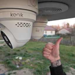 ASTA.PL SPÓŁKA Z OGRANICZONĄ ODPOWIEDZIALNOŚCIĄ - Dwie białe kamery Kenik zamontowane pod sufitem, z dłonią z tatuażem pokazującą kciuk w górę. W tle dom i zieleń. Nietypowy kadr z bliska.