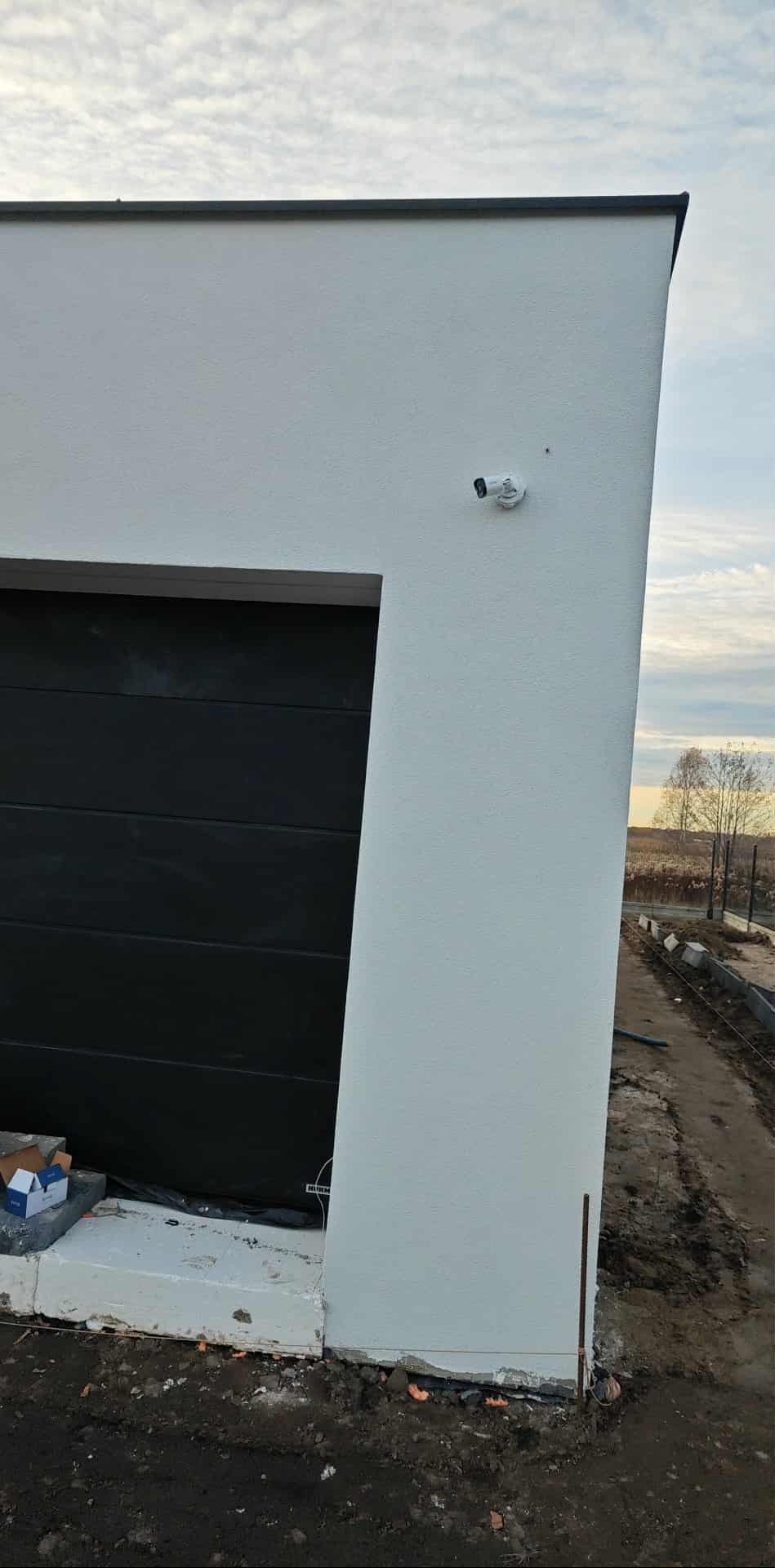 Biała kamera monitoringu na elewacji domu obok garażu z czarną bramą. Widok z dołu, na zewnątrz, w tle pole i niebo.