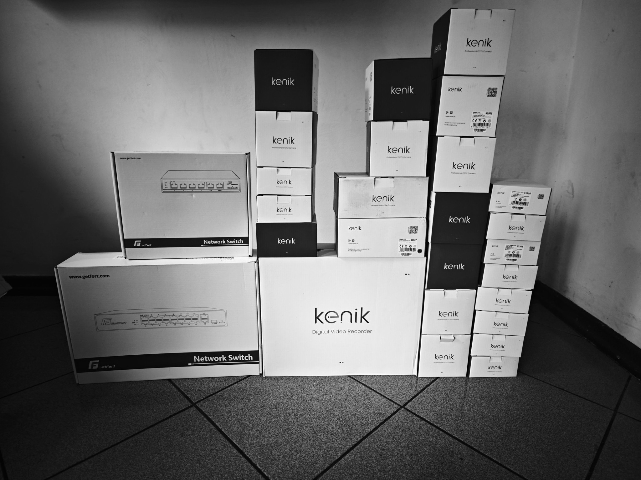 Stos pudełek Kenik (kamery CCTV, rejestratory) i GetFort (switche sieciowe) w czerni i bieli. Produkty do monitoringu wizyjnego ułożone na podłodze.