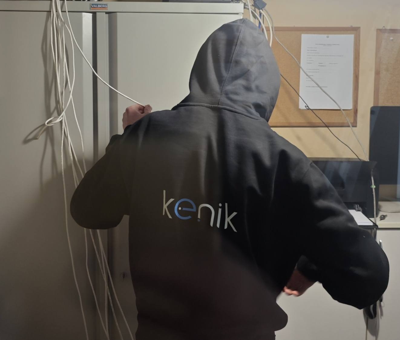 Osoba w bluzie z logo firmy Kenik, ciągnąca białe kable przy szafie Valberg. Widoczne dokumenty na korkowej tablicy i sprzęt biurowy w tle. Ujęcie od tyłu.