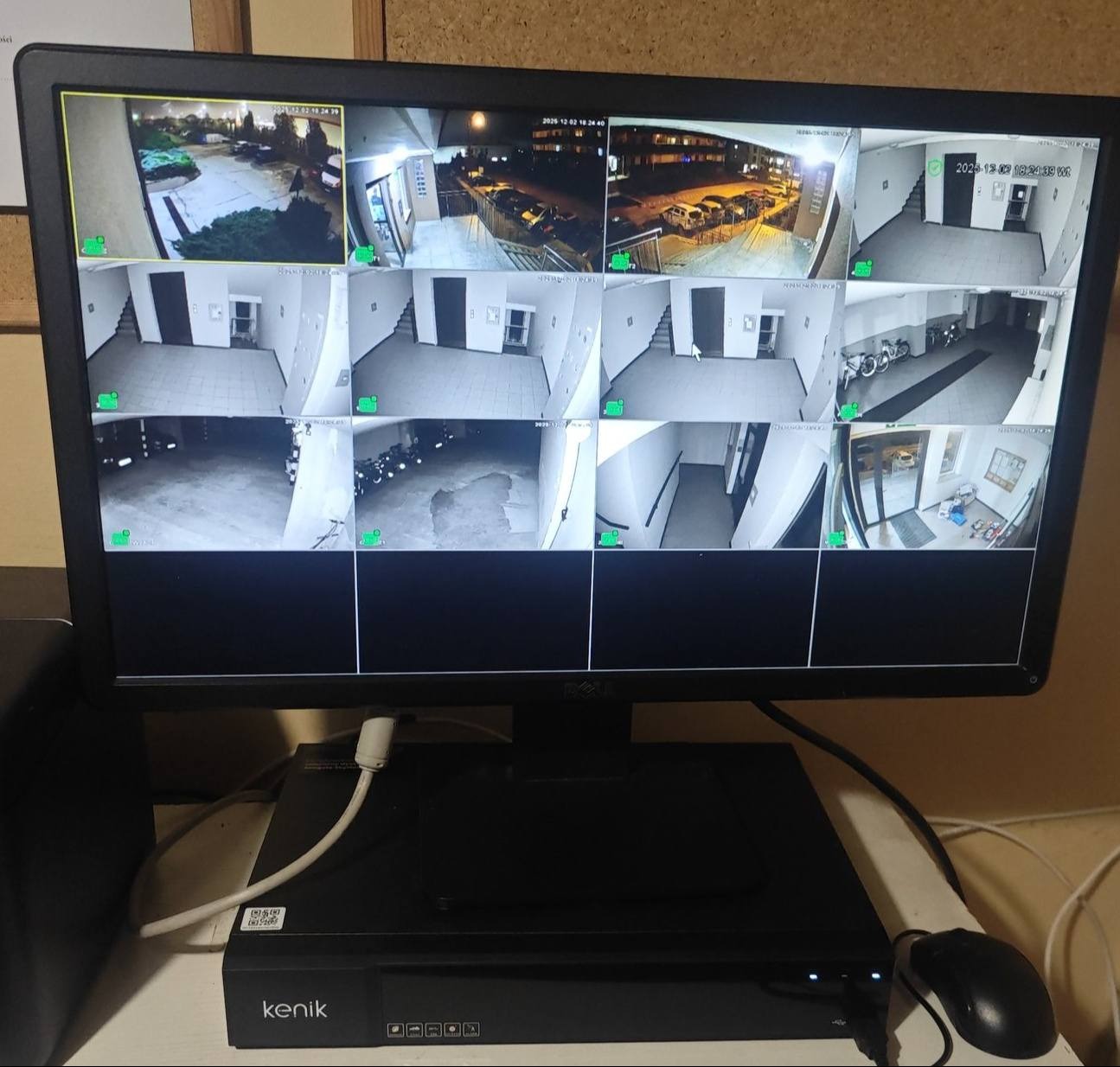 Ekran monitora z podglądem z kamer monitoringu Kenik, ukazujący różne obszary: parking, klatka schodowa, garaż. Widoczny rejestrator i mysz. Data i godzina na każdym z ujęć.