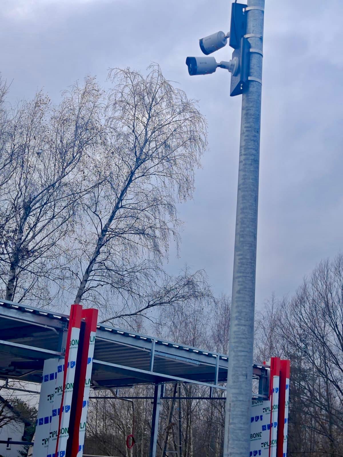 Dwie kamery monitoringu na słupie, widoczne z dołu, na tle pochmurnego nieba i drzew bez liści. W tle zadaszenie z elementami konstrukcyjnymi.