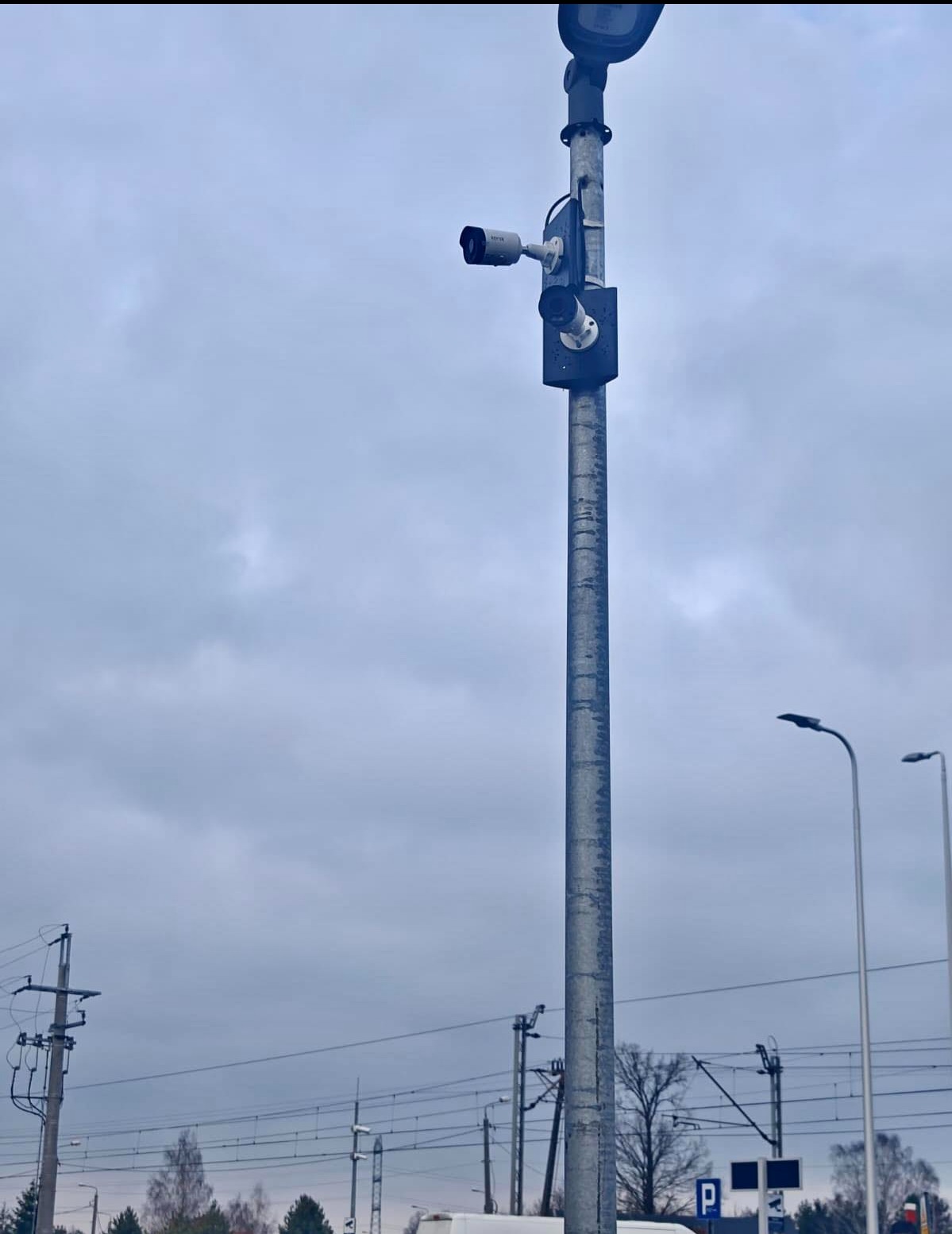 Słup z kamerami monitoringu na tle pochmurnego nieba, widoczne słupy energetyczne i oświetleniowe. Ujęcie z dołu, podkreślające wysokość instalacji.