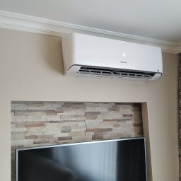 F.H.U. KLIMAKS Krzysztof Ślusarczyk - Biały klimatyzator Hisense zamontowany na ścianie nad telewizorem w salonie. Widoczne dekoracyjne wykończenie ściany z kamienia i wzorzyste zasłony.