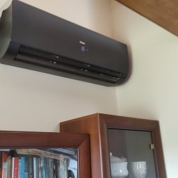F.H.U. KLIMAKS Krzysztof Ślusarczyk - Czarny klimatyzator Haier zamontowany na ścianie w jasnym wnętrzu, widoczny fragment drewnianej belki stropowej i górna część regału z książkami.