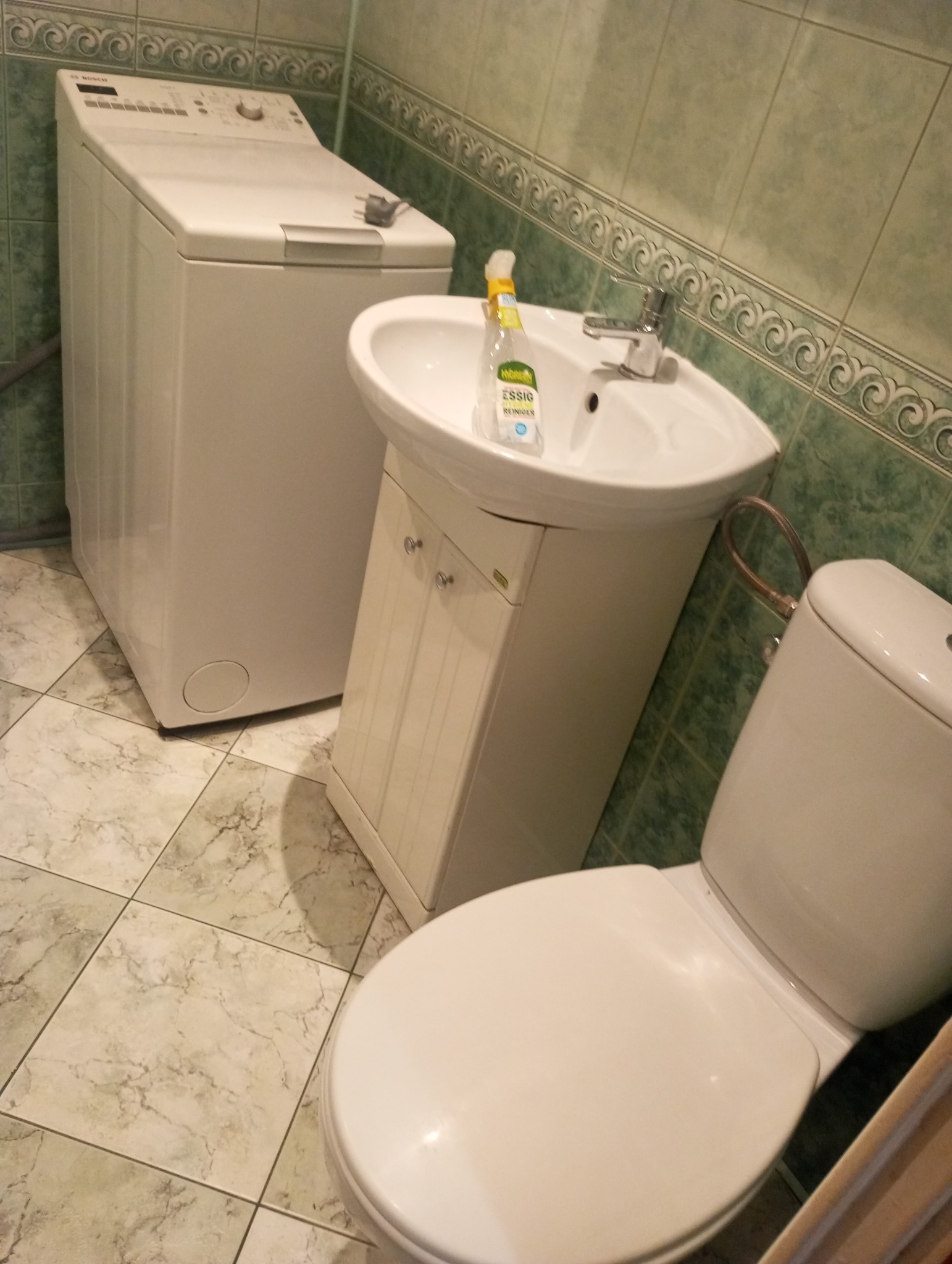 Wnętrze łazienki z białą pralką, umywalką z szafką i toaletą. Na umywalce stoi butelka środka czyszczącego. Ściany wyłożone zielonymi płytkami z dekoracyjnym wzorem.