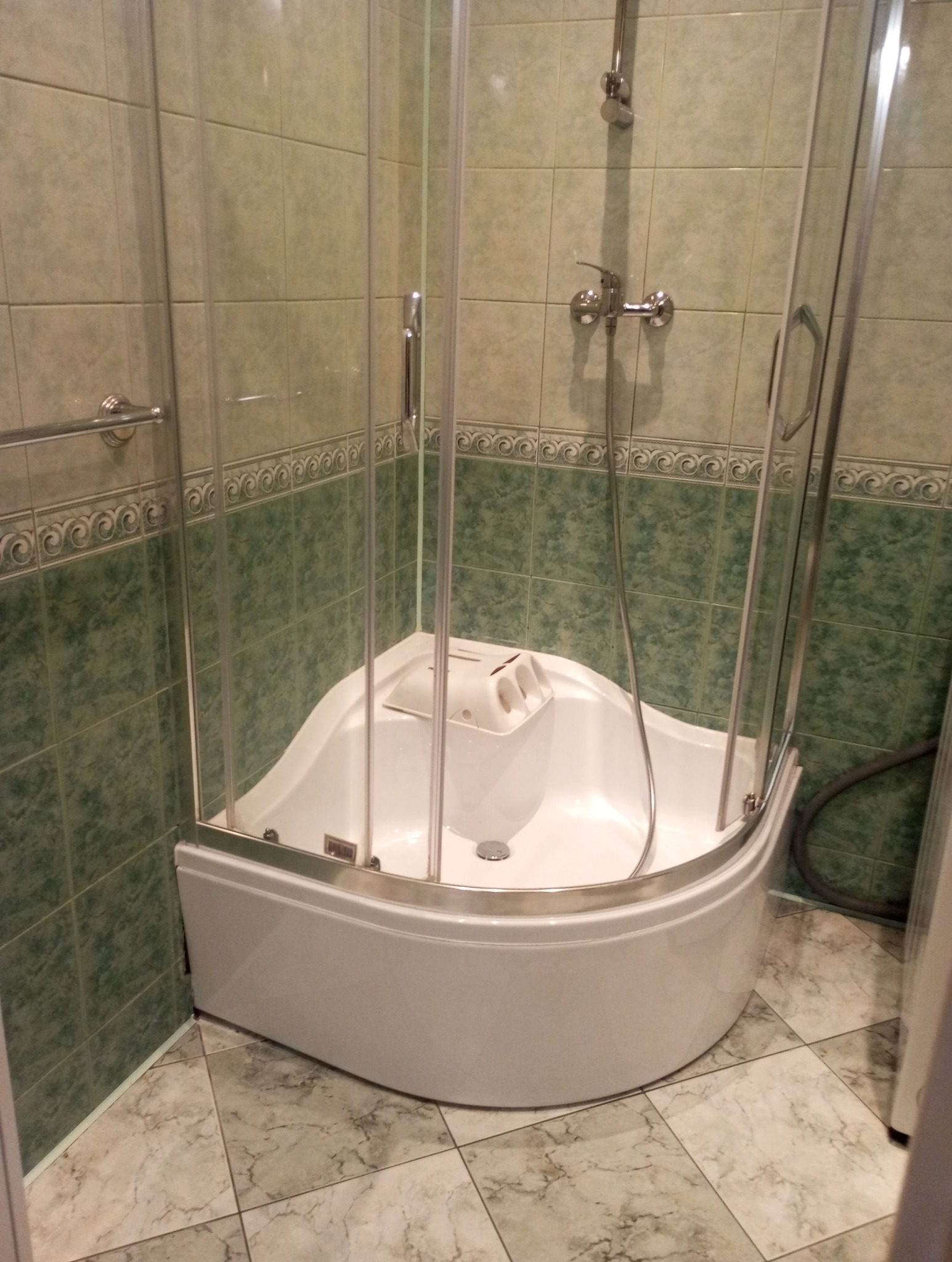 Wnętrze łazienki: narożna kabina prysznicowa z hydromasażem, biała, z przezroczystymi szybami, na tle zielonych płytek. Widok z bliska, bez osób.