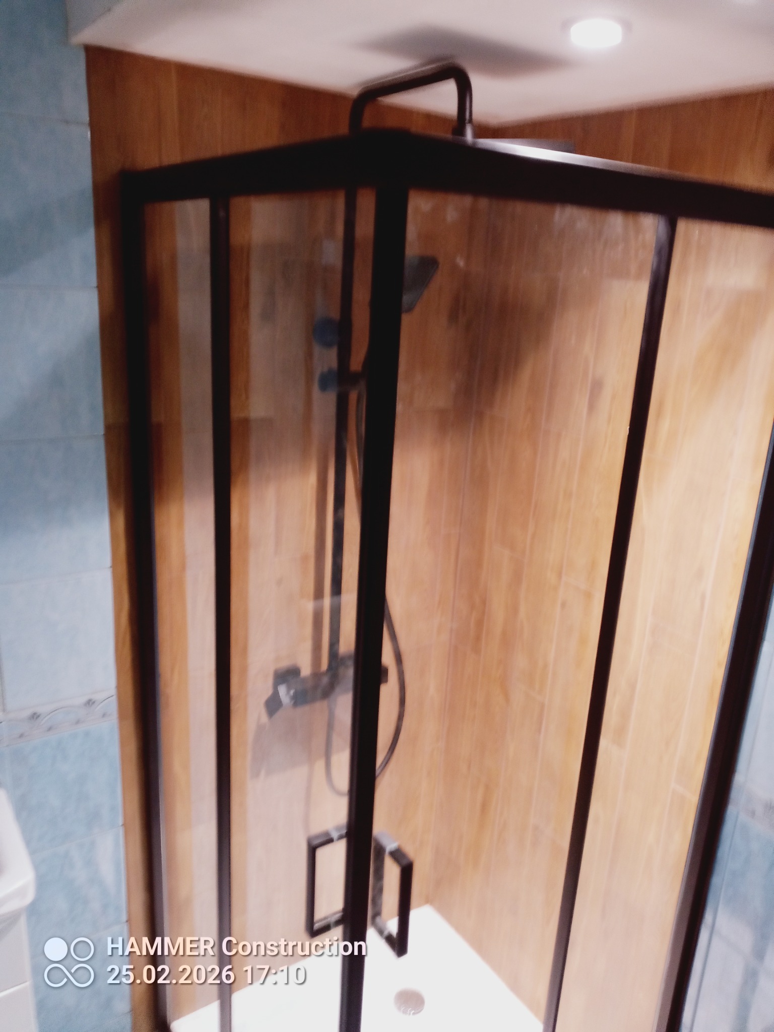 Nowoczesna kabina prysznicowa z czarnymi profilami i drewnopodobnymi płytkami na ścianach. Widoczny odpływ liniowy i bateria prysznicowa. Wykończenie łazienki.