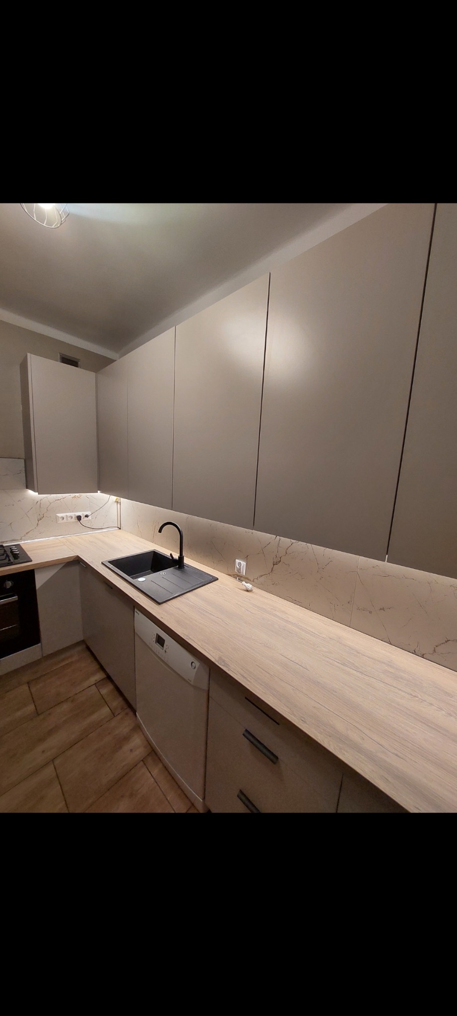 Beżowa kuchnia z drewnianym blatem, czarnym zlewem i marmurową ścianą. Minimalistyczny design z podświetleniem LED. Widoczne gniazdka elektryczne.