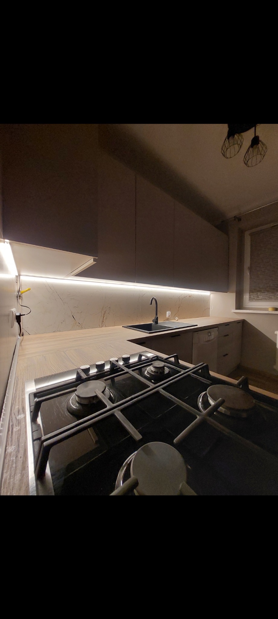 Nowoczesna kuchnia z szarymi szafkami, blatem z marmurowym wzorem i podświetleniem LED. Na pierwszym planie czarna płyta gazowa. Minimalistyczny design.