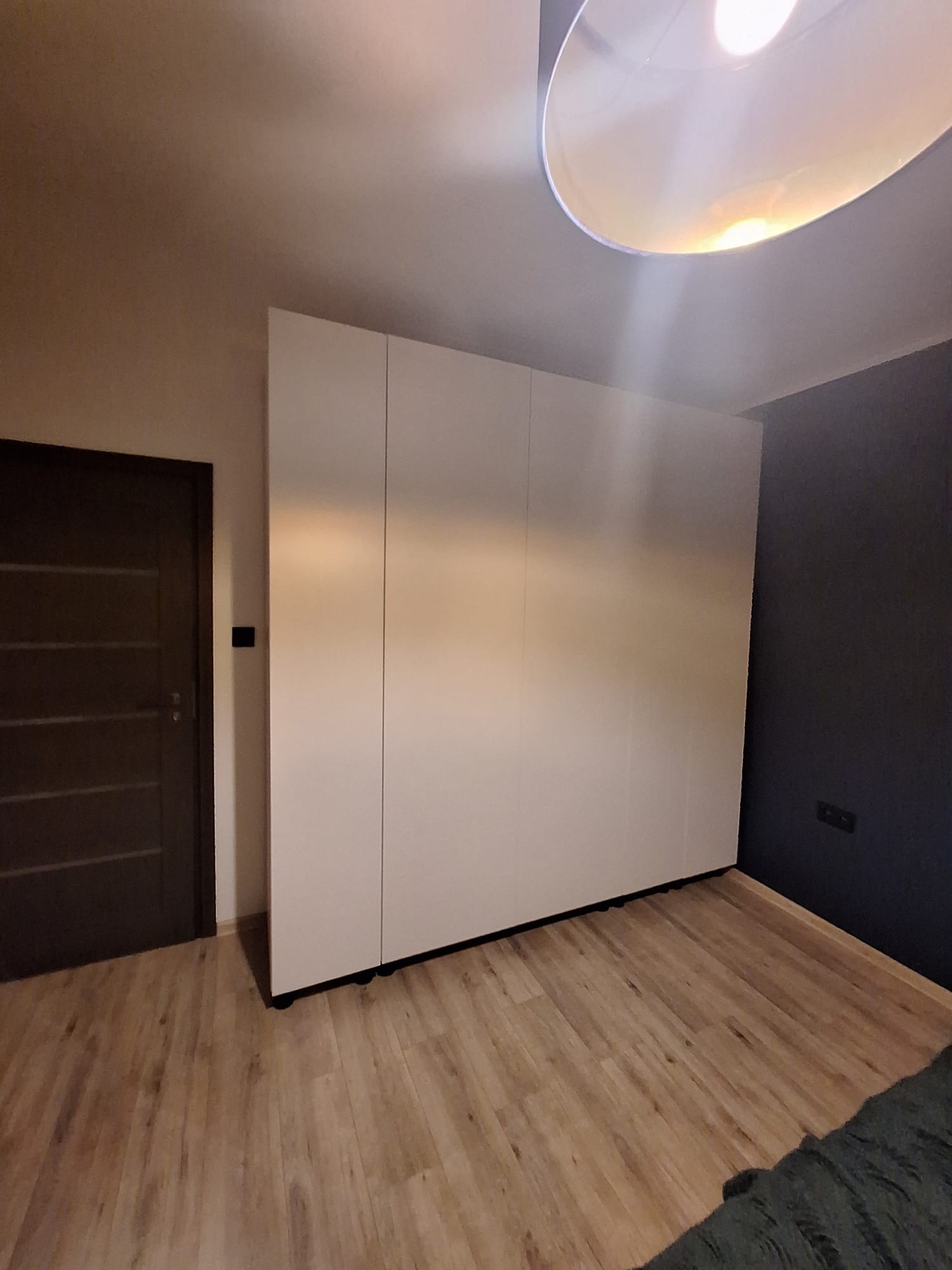 Biała, minimalistyczna szafa na wymiar z trzema drzwiami przesuwnymi, na tle drewnianej podłogi i ciemnej ściany. Widok z dołu, oświetlenie sufitowe.