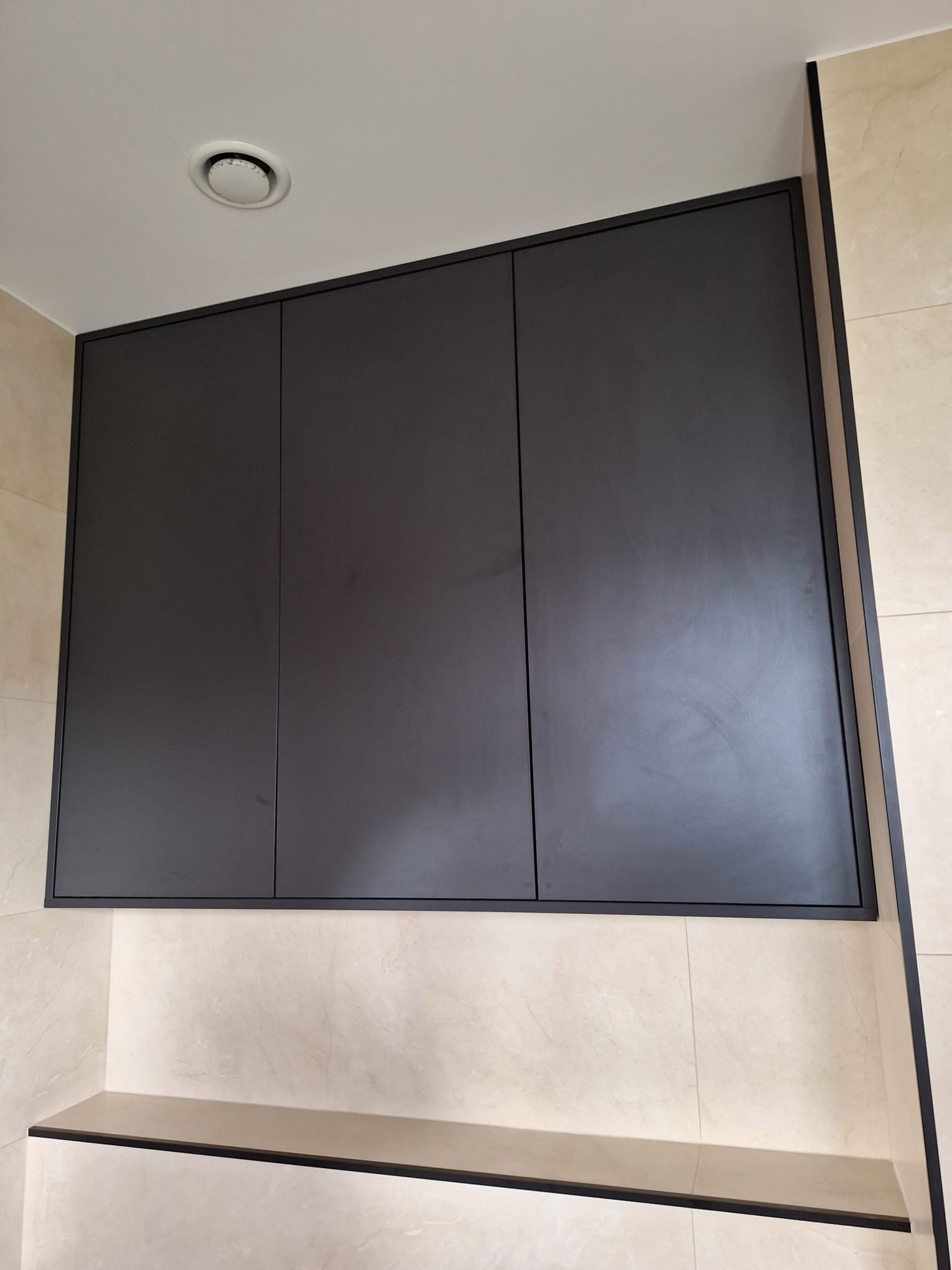 Nowoczesna, antracytowa szafka wisząca z trzema frontami, zamontowana nad półką w łazience wyłożonej płytkami imitującymi marmur.