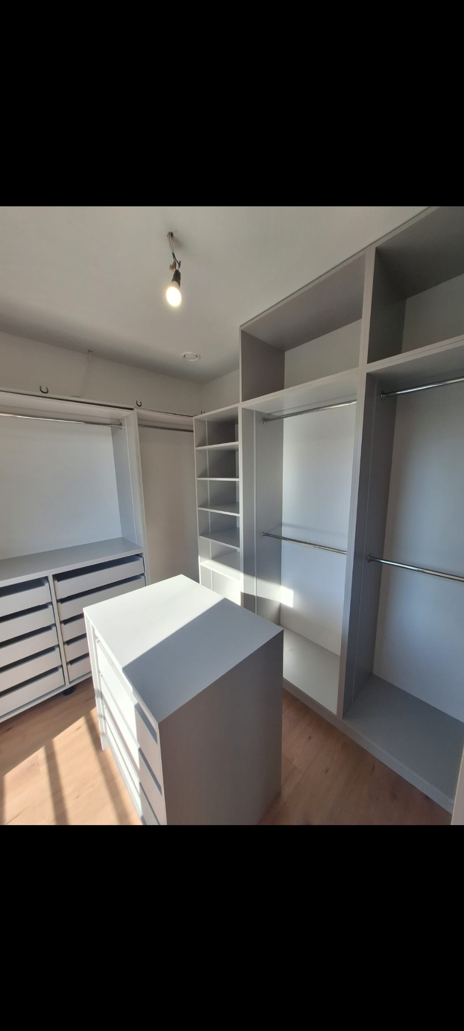 Jasna, przestronna garderoba z szufladami i półkami, oświetlona punktowym światłem, z drewnianą podłogą i widocznym światłem słonecznym.