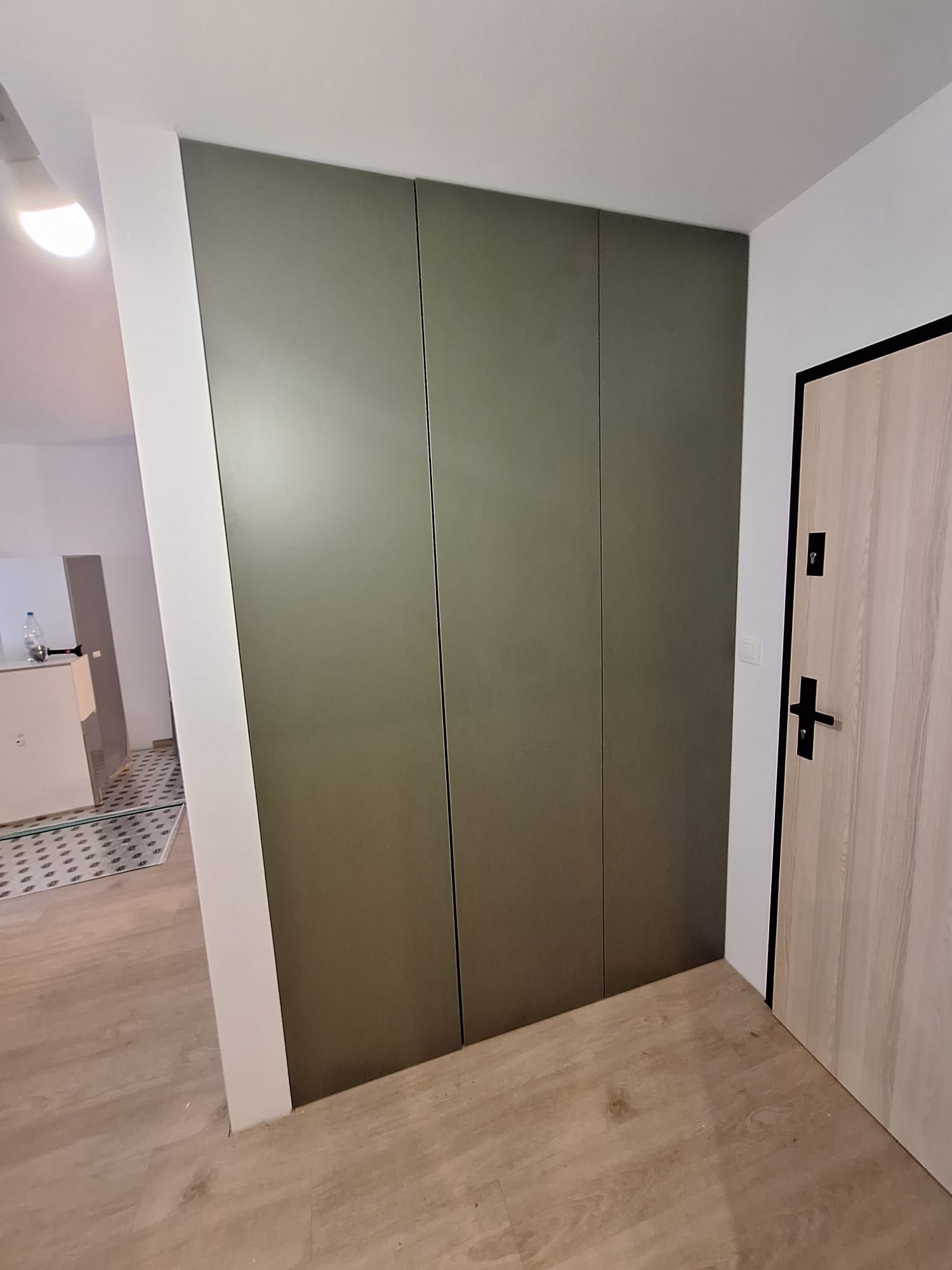 Nowoczesna, oliwkowa szafa wnękowa z trzema frontami, zlicowana ze ścianą, w minimalistycznym przedpokoju z drewnopodobną podłogą i jasnymi ścianami.