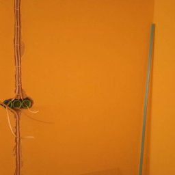 Elektro System - Żółta ściana z wyżłobieniami pod instalację elektryczną i puszkami. Widoczne przewody. Na podłodze obraz w ramie i inne przedmioty.