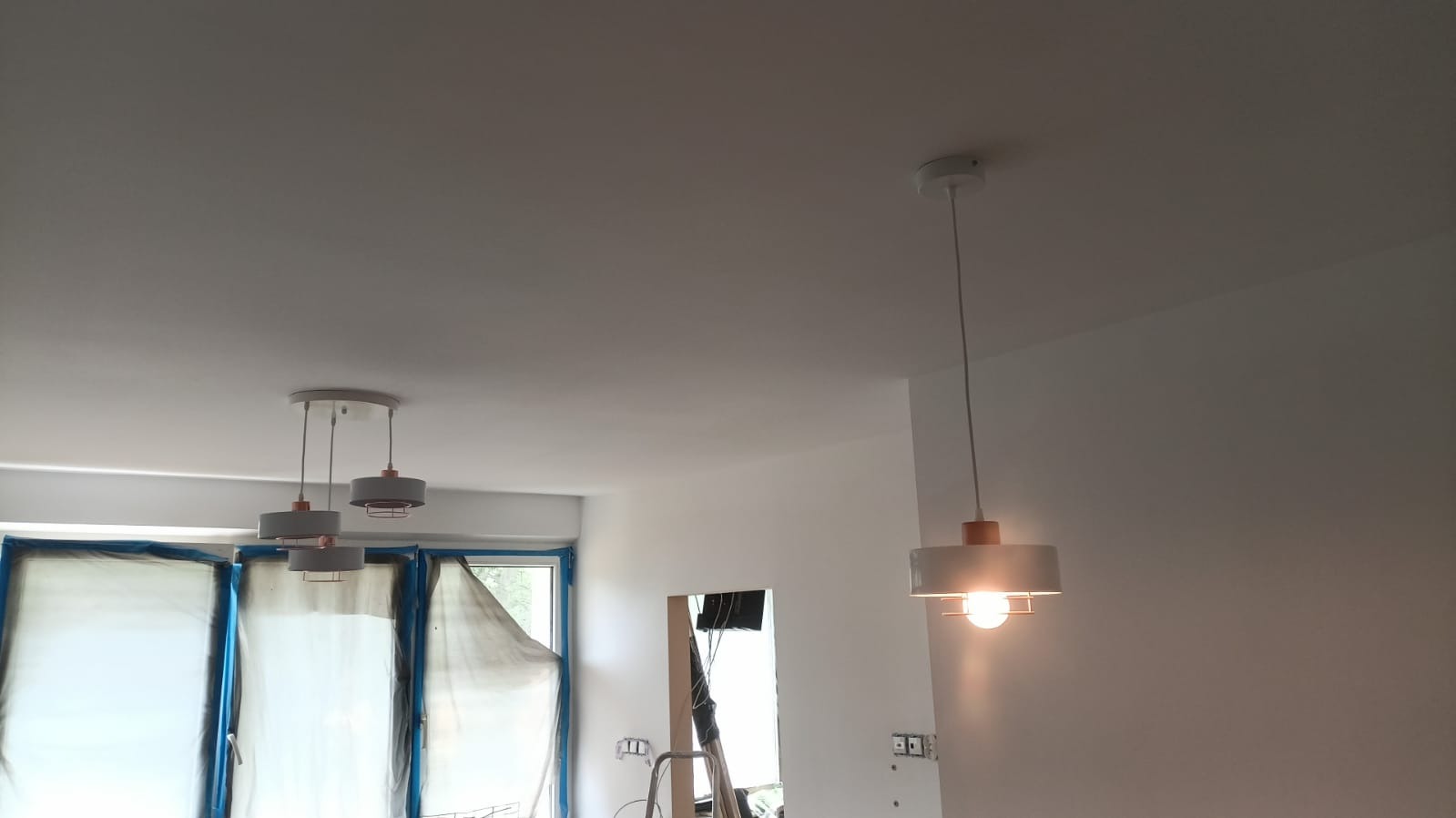Nowoczesne lampy wiszące w świeżo odmalowanym wnętrzu, okna zabezpieczone folią malarską, widoczne ślady prac wykończeniowych.
