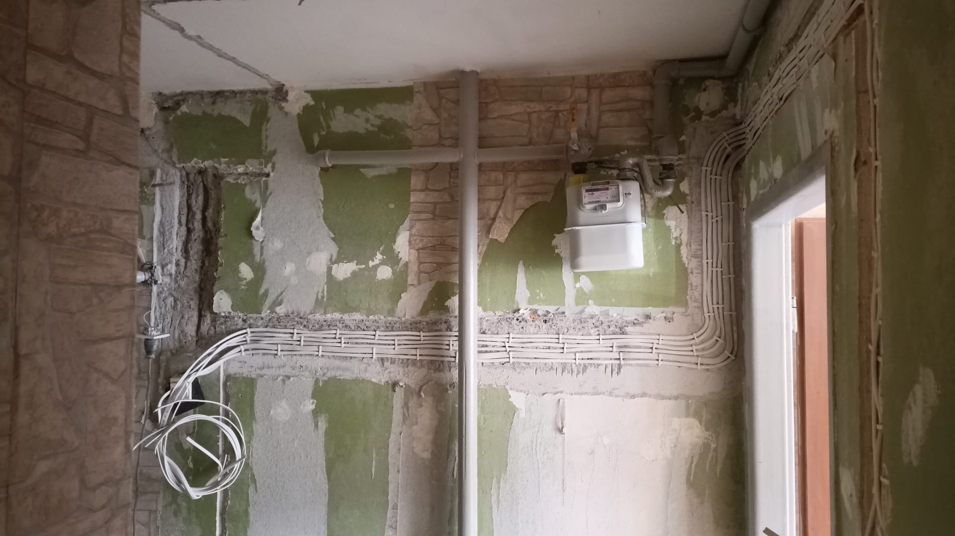 Instalacja elektryczna w trakcie remontu: białe przewody ułożone w bruzdach na ścianie, licznik gazu, surowe ściany z resztkami farby i tapet.
