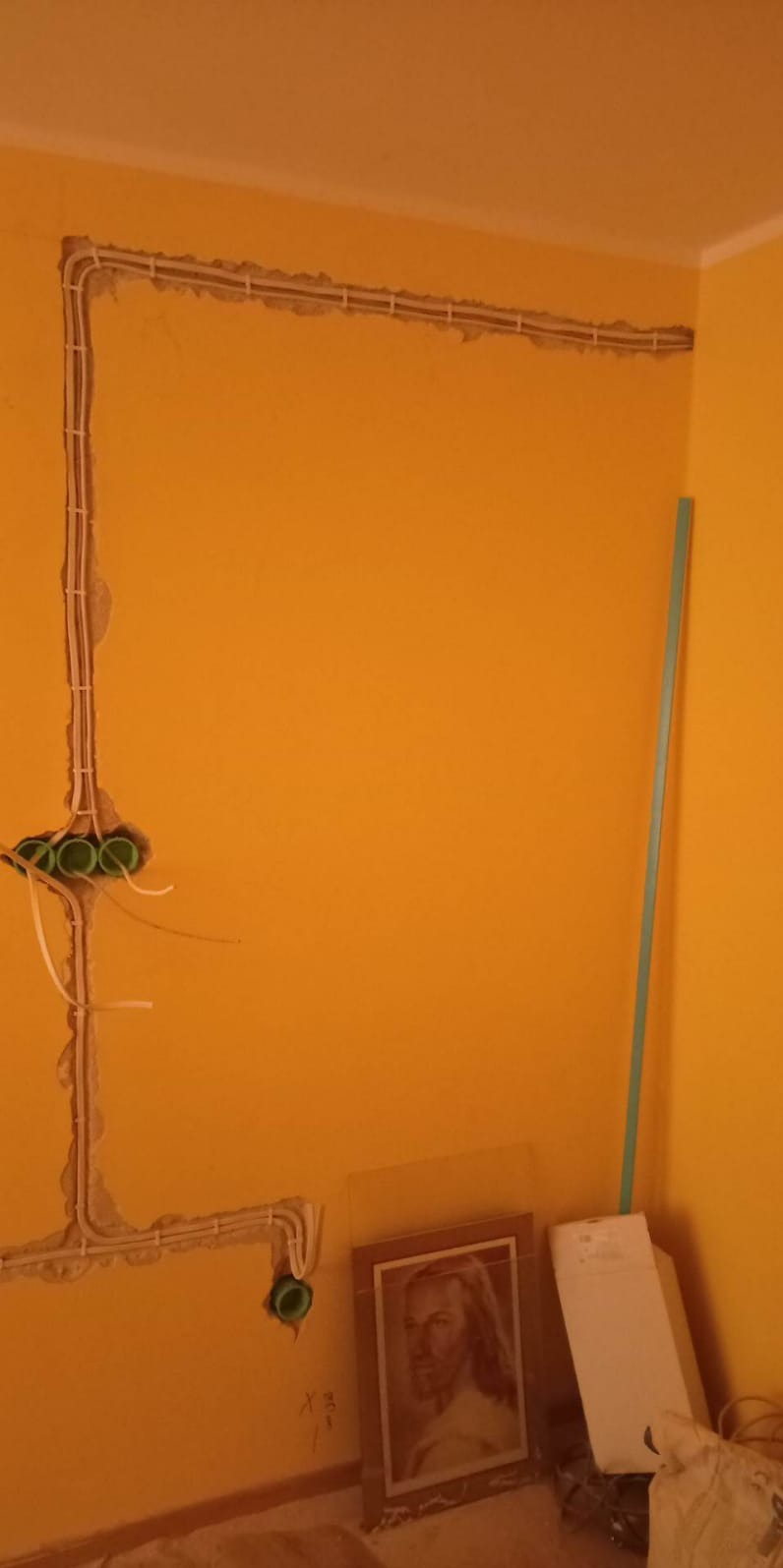 Żółta ściana z wyżłobieniami pod instalację elektryczną i puszkami. Widoczne przewody. Na podłodze obraz w ramie i inne przedmioty.