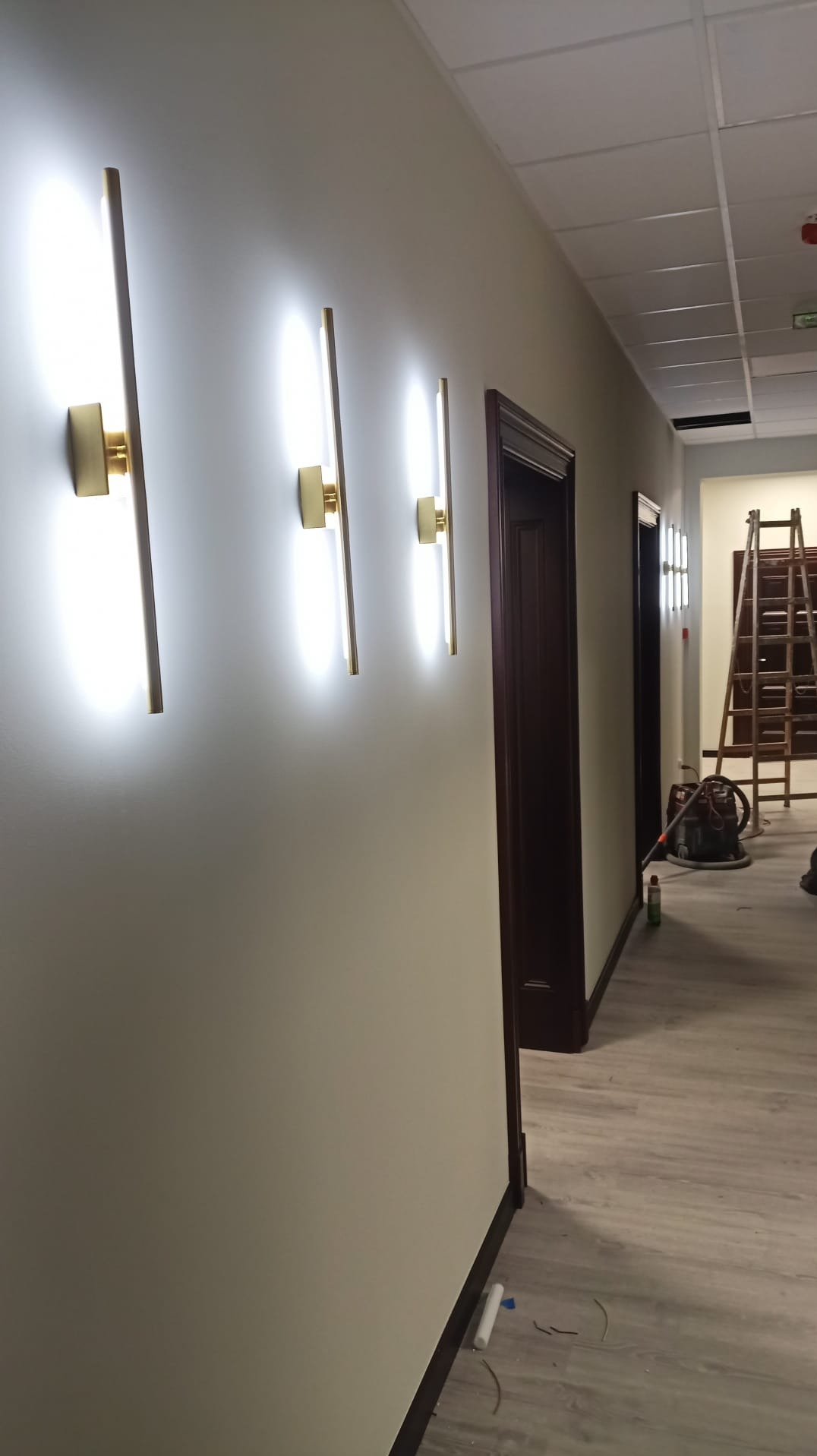 Korytarz z trzema nowoczesnymi, pionowymi lampami ściennymi o złotych akcentach, emitującymi jasne światło. W tle drzwi i drabina. Widok z boku.