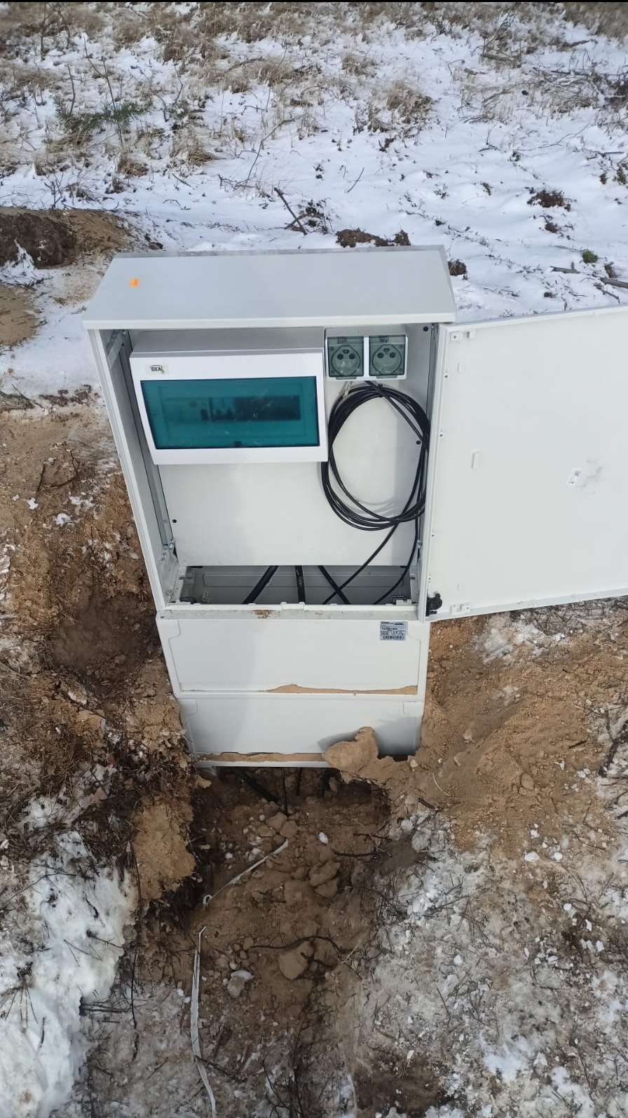 Otwarta, biała skrzynka elektryczna częściowo zagłębiona w ziemi na zewnątrz, z widocznymi kablami i elementami instalacji w środku. Zimowy krajobraz w tle.