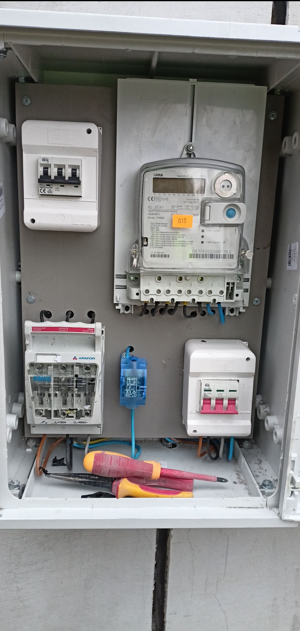 Otwarta skrzynka elektryczna z zamontowanymi bezpiecznikami, licznikiem energii i narzędziami wewnątrz. Widoczne przewody i elementy instalacji elektrycznej.