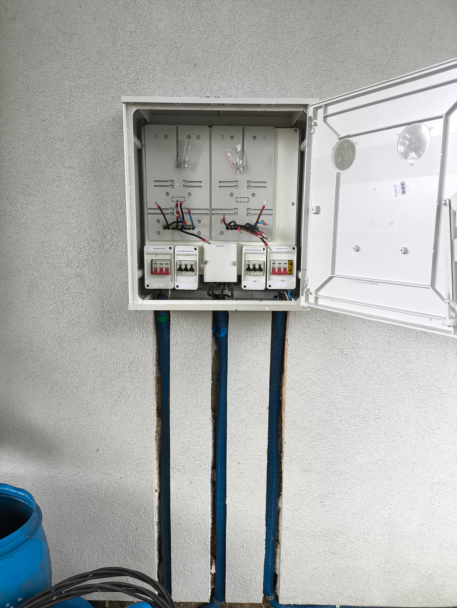 Otwarta skrzynka elektryczna z czterema licznikami i przewodami w ścianie o szorstkiej fakturze. Widoczne niebieskie rury instalacyjne.