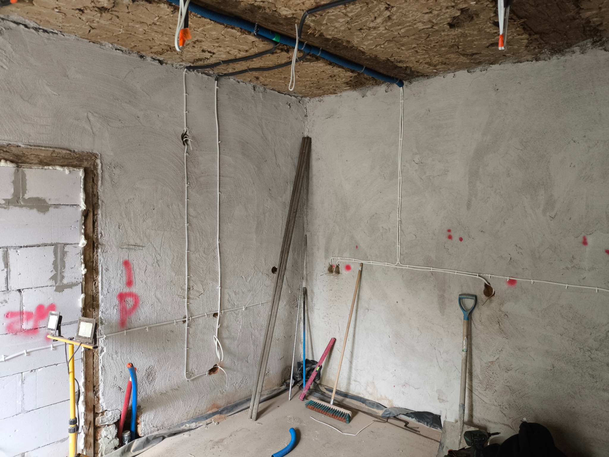 Surowy pokój w trakcie prac wykończeniowych: instalacja elektryczna w białych kablach, nieotynkowane ściany z czerwonymi oznaczeniami, narzędzia budowlane.