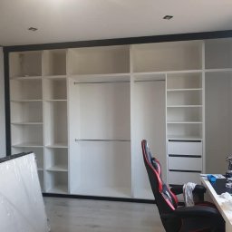 Meble Mirocha - Biała szafa wnękowa z czarną ramą w pokoju w Kalwarii Zebrzydowskiej. Widoczne półki, drążki i biurko z krzesłem gamingowym. Częściowo zapakowane elementy szafy.