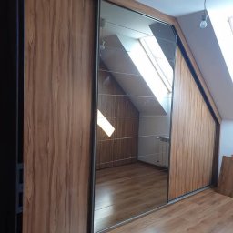 Meble Mirocha - Szafa wnękowa z lustrzanymi drzwiami przesuwnymi, drewnopodobne wykończenie, dopasowana do skosu poddasza, widoczne odbicie okna dachowego.
