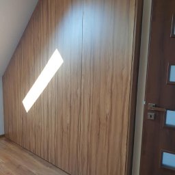 Meble Mirocha - Drewniana szafa wnękowa na wymiar, dopasowana do skosu poddasza. Widoczny fragment drzwi po prawej stronie. Naturalne światło podkreśla fakturę drewna.