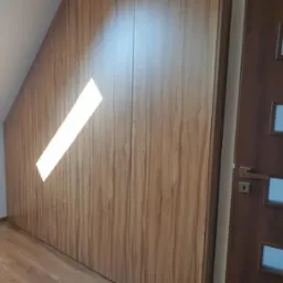 Drewniana szafa wnękowa na wymiar, dopasowana do skosu poddasza. Widoczny fragment drzwi po prawej stronie. Naturalne światło podkreśla fakturę drewna.
