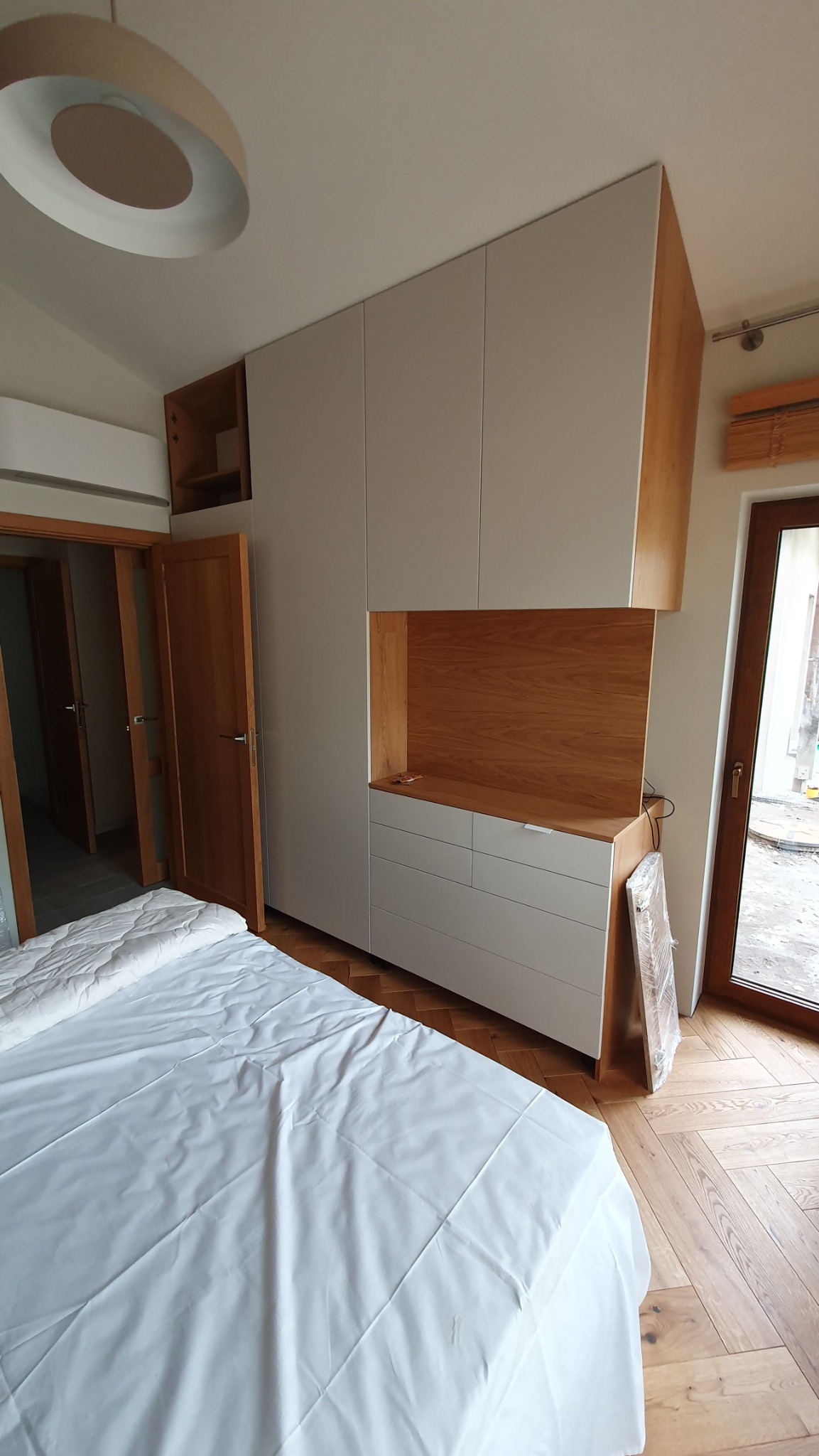 Szafa na wymiar w sypialni: zabudowa z białymi frontami i drewnianymi akcentami, widok na łóżko z białą pościelą i parkiet w jodełkę, nowoczesny design.