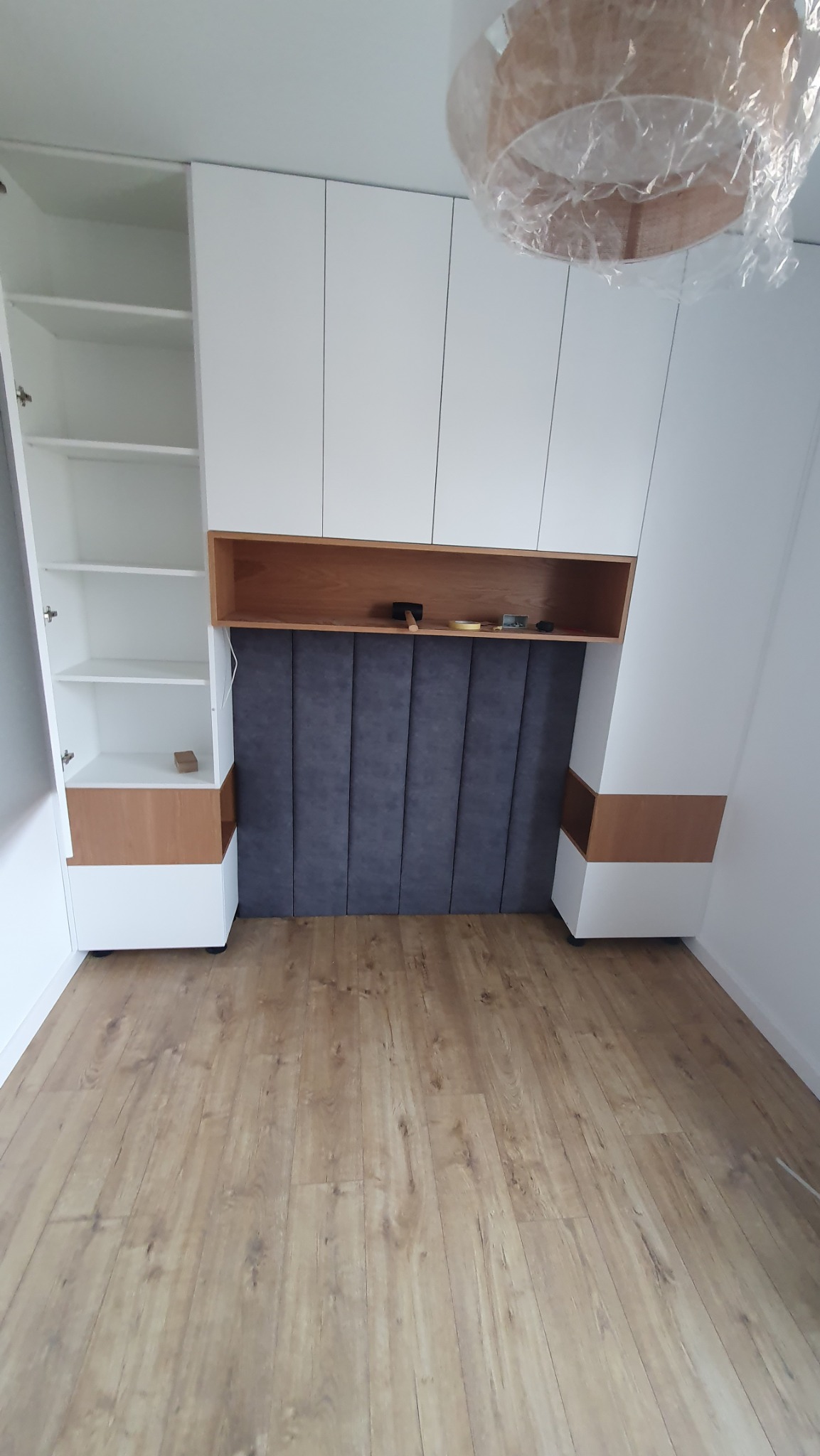 Biała szafa z otwartą półką i drewnianymi akcentami, nad tapicerowanym wezgłowiem łóżka. Na półce leżą narzędzia. Nowoczesny design, jasne wnętrze.