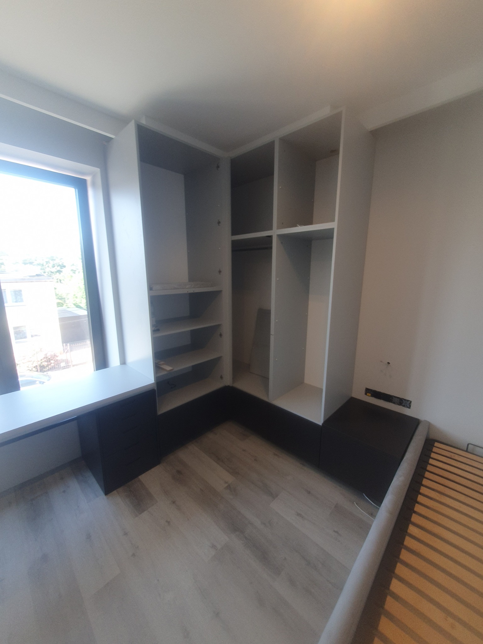 Jasna szafa wnękowa z półkami i drążkiem, wkomponowana w przestrzeń pokoju, zintegrowana z biurkiem i elementami łóżka, widok na okno.